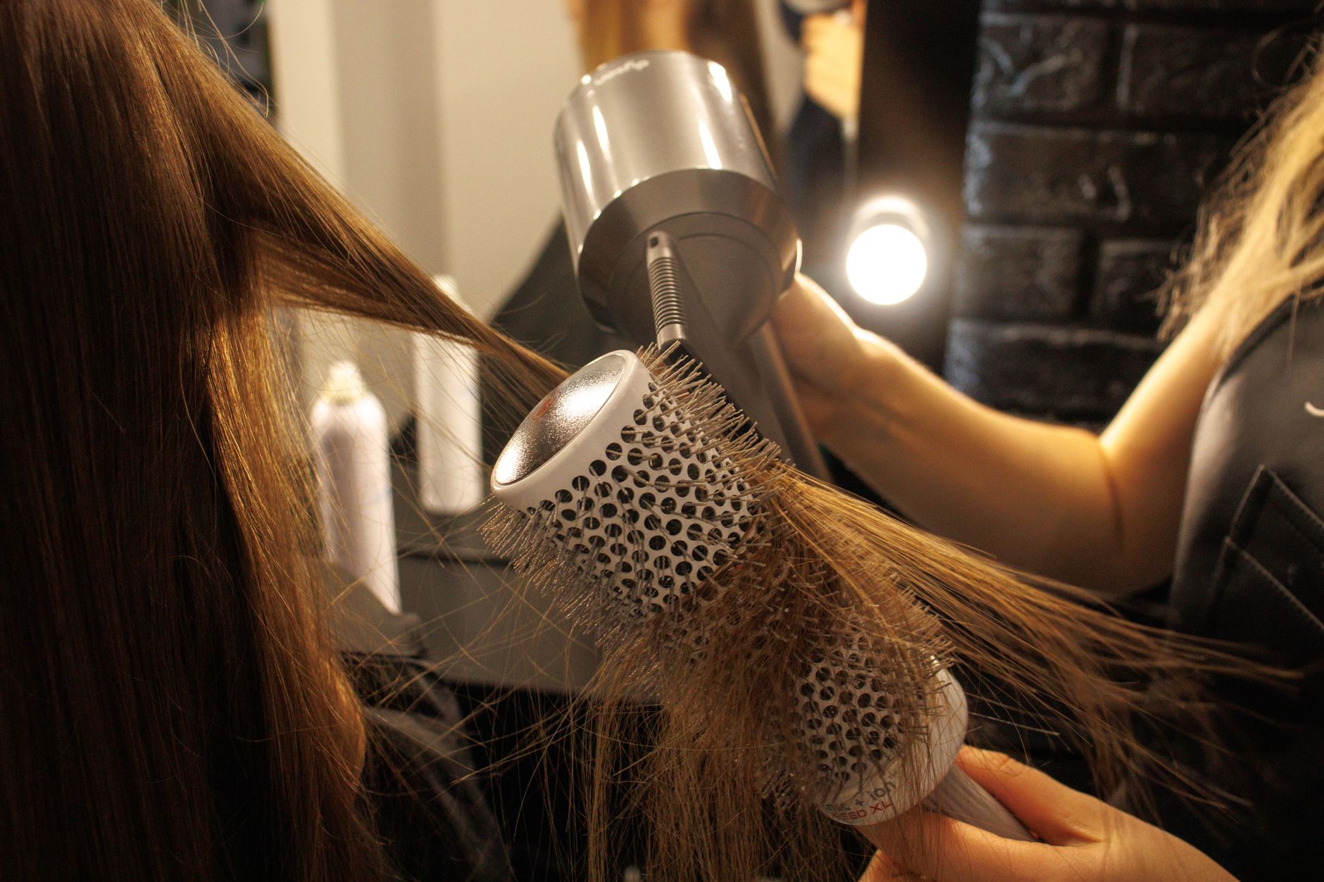 A stylist smoothing long, glossy hair with a round brush under warm salon light